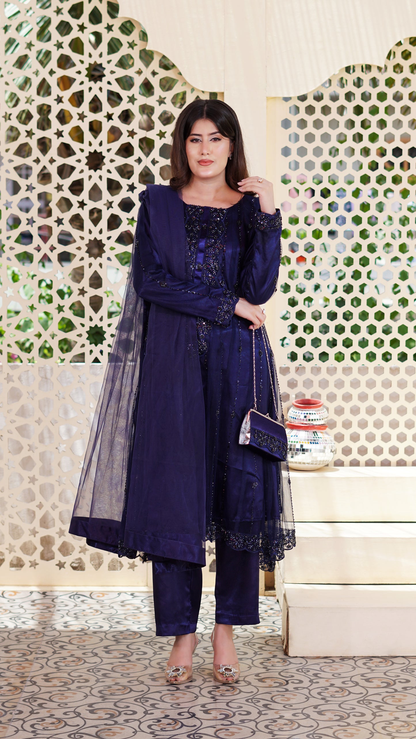 A woman models a deep navy blue ethnic three-piece suit with matching sheer dupatta, embellished with intricate embroidery and lace. She is holding a matching embroidered clutch bag and posing in front of a decorative white screen with geometric patterns.