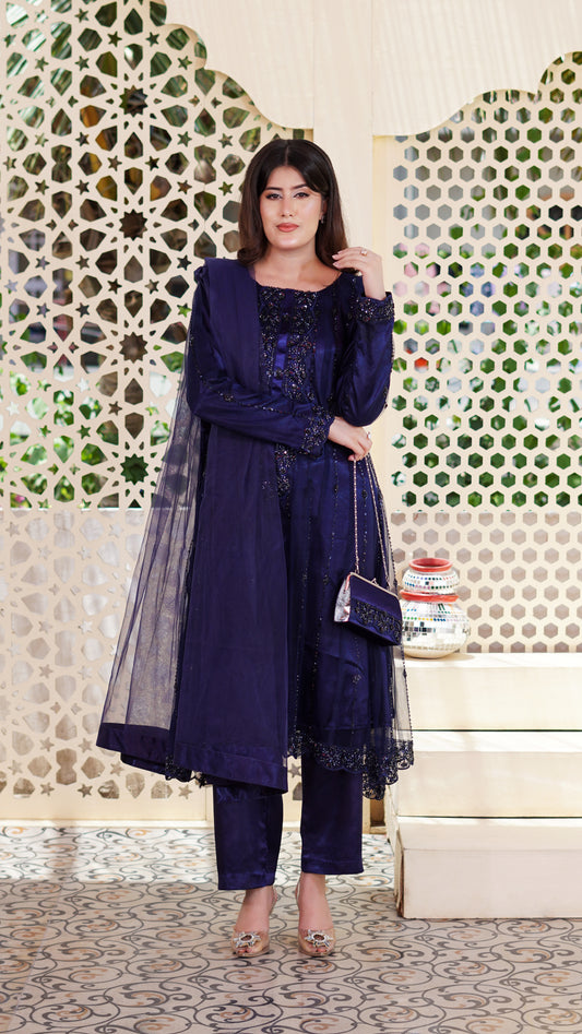 A woman models a deep navy blue ethnic three-piece suit with matching sheer dupatta, embellished with intricate embroidery and lace. She is holding a matching embroidered clutch bag and posing in front of a decorative white screen with geometric patterns.