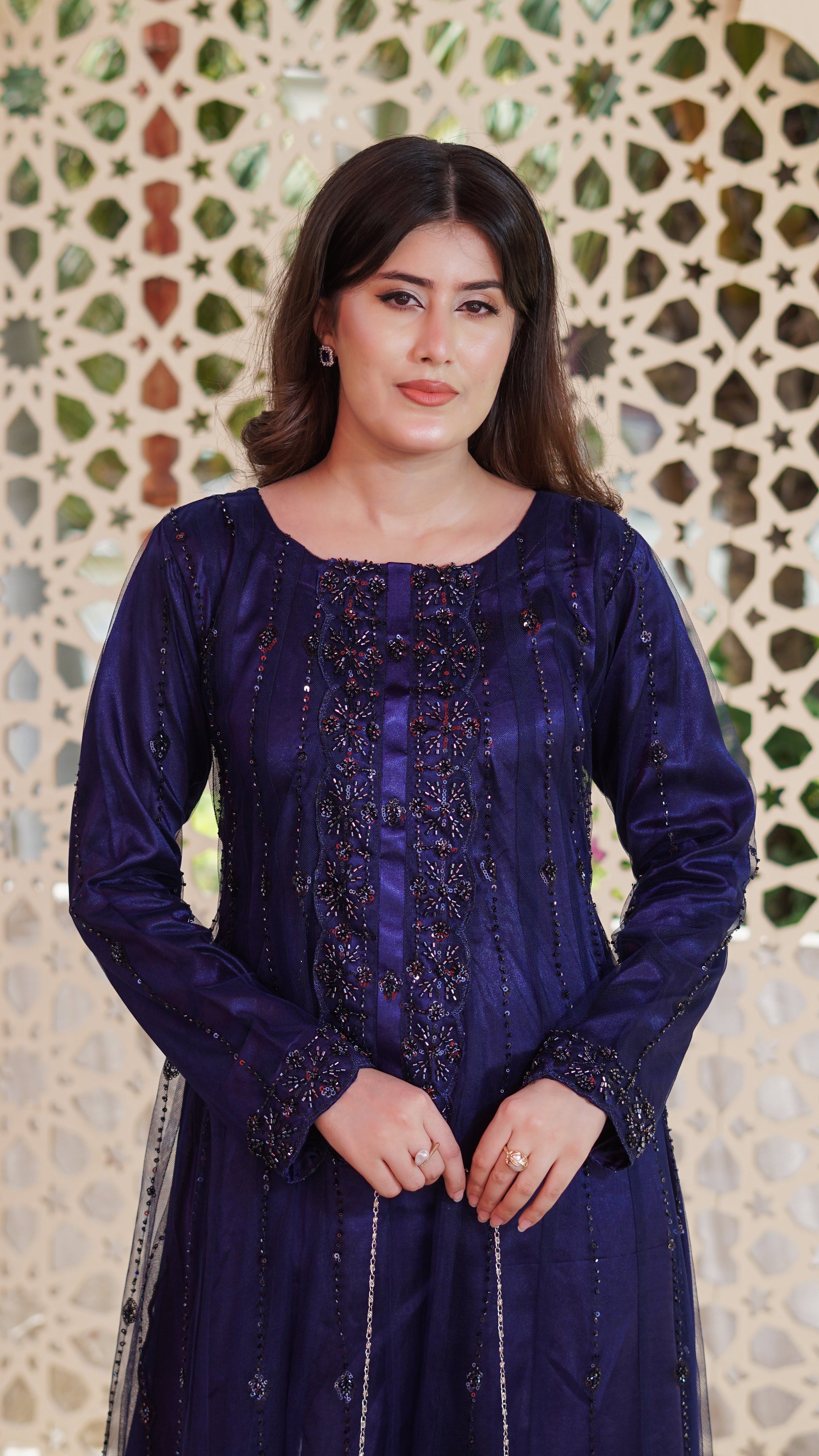 A woman wearing a deep purple embroidered traditional outfit with long sleeves and intricate beadwork, standing in front of a decorative geometric patterned wall.